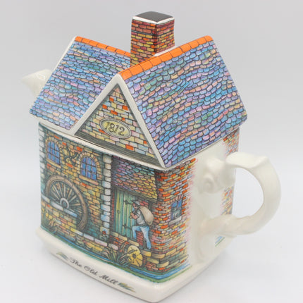 A ceramic teapot shaped like a detailed house, featuring a colorful tiled roof, brick walls, a water wheel illustration, and a white handle on a light gray background.