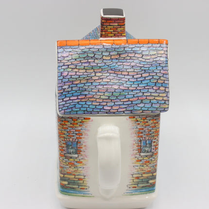 A ceramic house-shaped mug with a colorful painted roof and brick-textured walls, featuring a white handle and a small chimney detail on top, photographed against a light gray background.