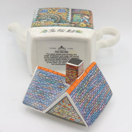 A ceramic tea towel shaped like a house, shown both upright with its printed front design visible and lying on its side to display the detailed back print and handle. The front features a colorful mosaic-style roof design in blue, orange, and white, with a small brick chimney.