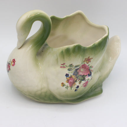 A decorative ceramic pitcher shaped like a swan, featuring a cream-colored body with a green neck and tail, and a floral pattern with pink, red, and blue blossoms on the side.