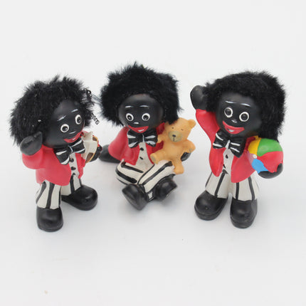 Three black-and-white Golly figurines with black hair, red jackets, and striped pants, posed together on a plain white background. One holds a small tan teddy bear, and one holds a multicolored ball.