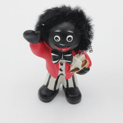 A small figurine of a Golly with black skin, black hair, and a black-and-white striped outfit with a red jacket, holding a white object, photographed against a plain white background.