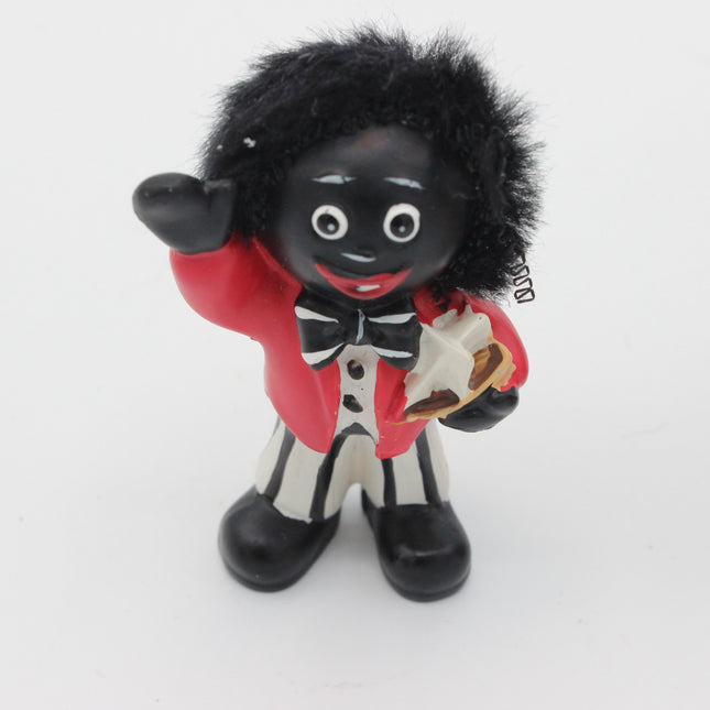 A small figurine of a Golly with black skin, black hair, and a black-and-white striped outfit with a red jacket, holding a white object, photographed against a plain white background.