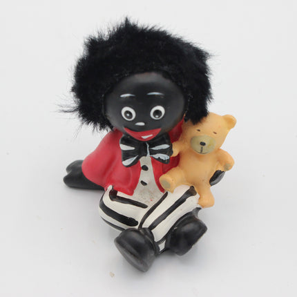 A small figurine of a black-skinned Golly character with black hair, wearing a red jacket, white shirt, black bow tie, and striped pants, while holding a tan teddy bear, photographed on a plain white background.