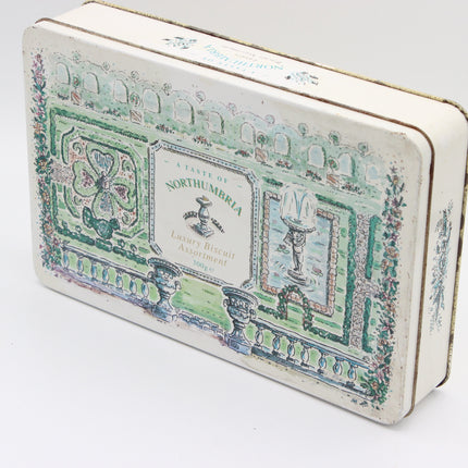 A vintage rectangular metal biscuit tin with an illustrated Northumbrian garden design in green, blue, and pink on an off-white background, photographed on a white studio background.