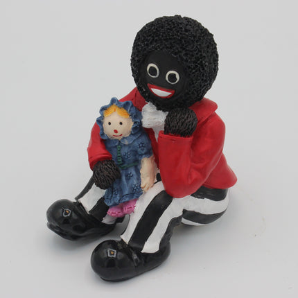 A collectible Golly figurine of a black-and-white character wearing a red jacket and holding a small blonde doll, photographed against a plain light gray background.