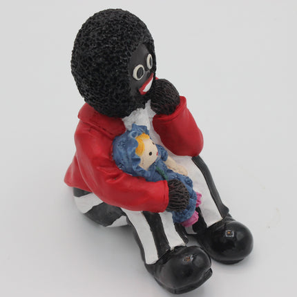A glossy black-and-white Golly doll figurine wearing a red jacket and holding a small blue-and-white stuffed doll, photographed on a plain light gray background.