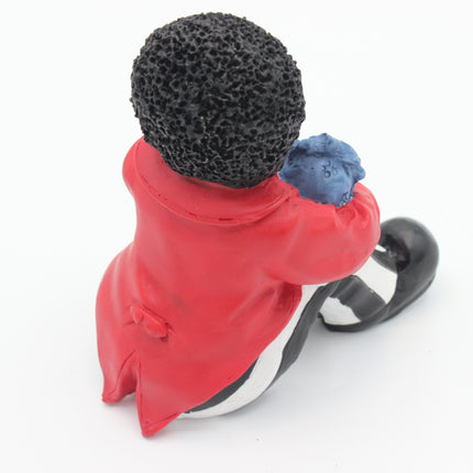 A small figurine of a Golly seen from behind, wearing a bright red coat, black-and-white striped pants, and a black textured hat, holding a blue object, photographed on a white background.
