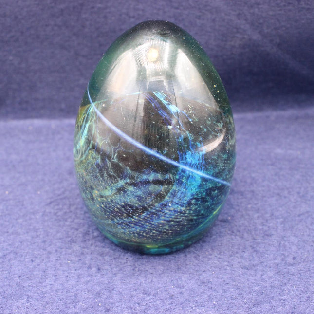 A polished glass paperweight with swirling blue, green, and black patterns, set against a textured purple background.