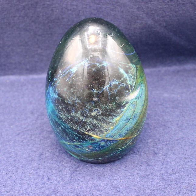 A polished glass paperweight with swirling blue, green, and black patterns, resting on a textured purple fabric background.