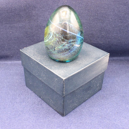 A polished iridescent glass paperweight with swirling blue, green, and yellow tones, displayed on top of a dark gray textured box against a purple fabric background.