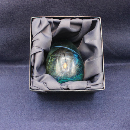 A polished iridescent green and blue paperweight nestled inside a square gray gift box lined with gray fabric, set against a dark blue background.