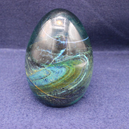 A polished glass paperweight featuring a swirling cosmic pattern of teal, green, blue, and gold, set against a textured purple background.