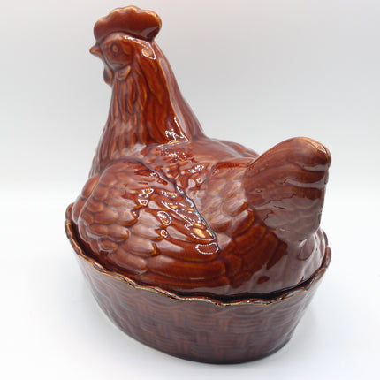 A glossy brown glazed ceramic lidded container shaped like a rooster standing over a basket-like base, photographed on a plain light gray studio background.