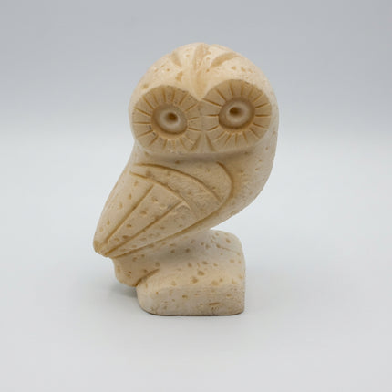 A small carved owl figurine in a light cream or ivory tone, photographed against a plain light gray studio background.