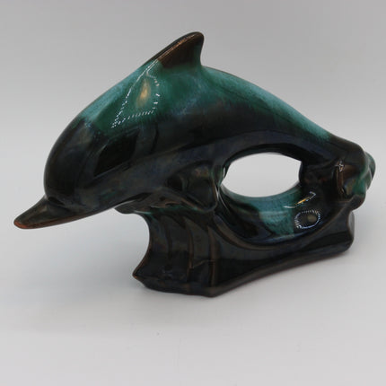 A sculptural ceramic dolphin figurine with a glossy glaze, featuring a color transition from deep black to teal green, photographed on a neutral light gray background.