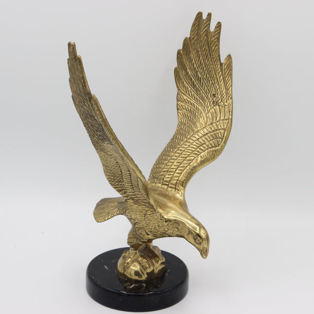 A metallic gold eagle sculpture with wings spread, perched on a small golden ball and mounted on a round black base, photographed against a plain light gray studio background.