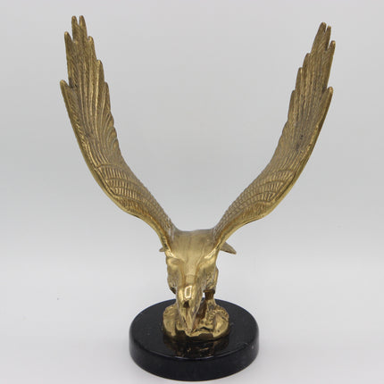 A metallic gold eagle skull sculpture with wings spread wide, mounted on a round black base against a light gray studio background.