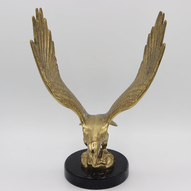 A metallic gold eagle skull sculpture with wings spread wide, mounted on a round black base against a light gray studio background.