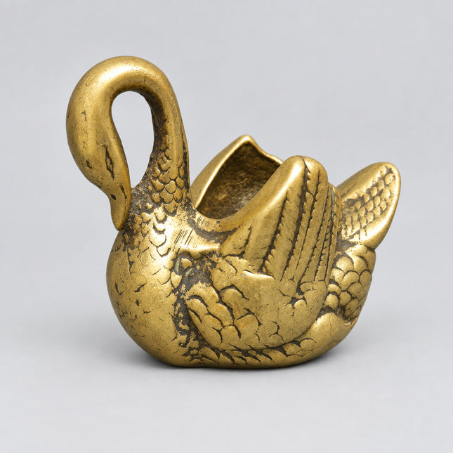 A small metallic swan-shaped object with a curved neck and open top, shown on a solid green background.