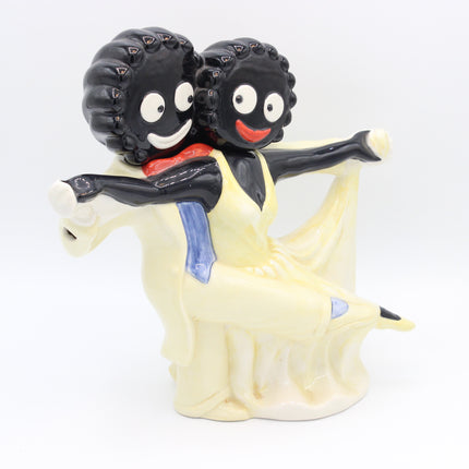 A ceramic figurine of two Gollies one with curly black hair and one with straight black hair, both smiling and wearing yellow dresses with blue sashes, posed against a plain white background.