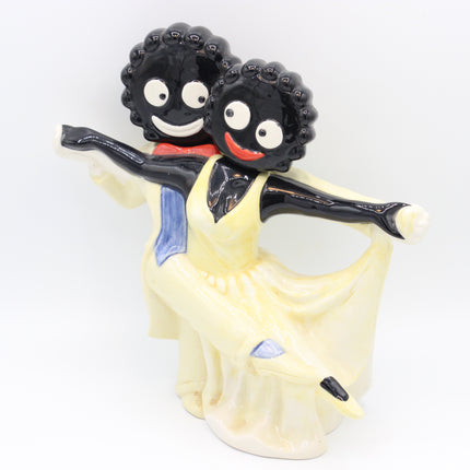 A ceramic figurine of two dancing figures, one with black skin and curly hair wearing a yellow dress and blue sash, and the other with black skin and curly hair wearing a red top, set against a plain white background.