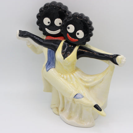 A glossy ceramic figurine of two dancing figures with black heads, white eyes, and wide smiles. One wears a pale yellow dress with a blue sash, and the other wears a red collar and blue sash, posed against a plain light gray background.