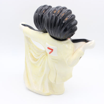 A ceramic figurine of a Golly in a cream-colored kimono with a small red symbol on her back, carrying a large black ribbed bundle on her head, photographed against a white background.
