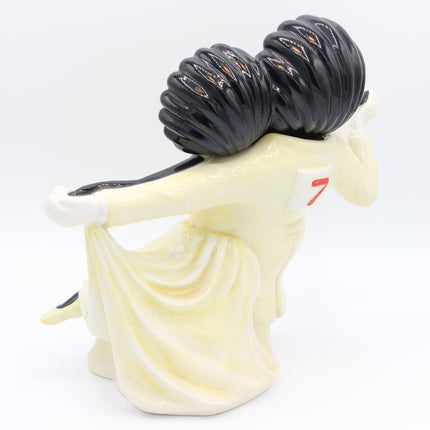 A glossy ceramic figurine of a Golly with a large black hair bun, wearing a cream-colored dress with a small red number 7 on the back, shown against a plain white background.