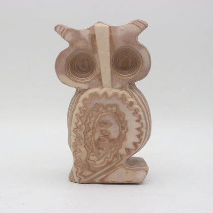 A carved stone owl figurine with concentric circular patterns in shades of beige, cream, and light brown, set against a plain light gray background.