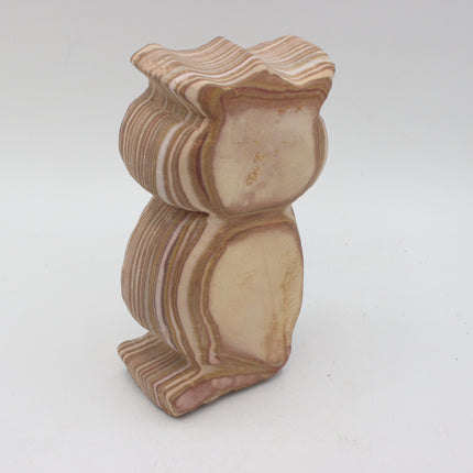 A carved stone owl figurine with natural layered banding in beige, cream, and light brown tones, photographed on a plain light gray background.