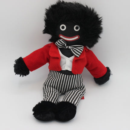 A plush Golly doll with black fuzzy hair and black feet, wearing a red jacket, white shirt, black-and-white striped bow tie and striped pants, photographed against a plain light gray background.