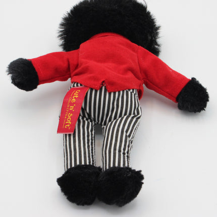 A small plush Golly doll or toy figure shown from the back, wearing a bright red top, black-and-white vertically striped pants, and black fuzzy trim at the neck, cuffs, and feet. A red fabric tag is attached to the back.