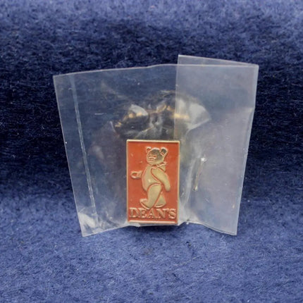 A small rectangular enamel pin with a gold bear design and the word 'DEANS' on an orange-red background, sealed inside a clear plastic protective bag and placed on a textured blue surface.
