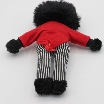 A small doll or plush Golly viewed from behind, wearing a bright red top, black-and-white vertically striped pants, and black fuzzy trim on the head, arms, and feet, photographed on a plain light background.
