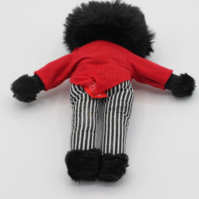 A small doll or plush Golly viewed from behind, wearing a bright red top, black-and-white vertically striped pants, and black fuzzy trim on the head, arms, and feet, photographed on a plain light background.