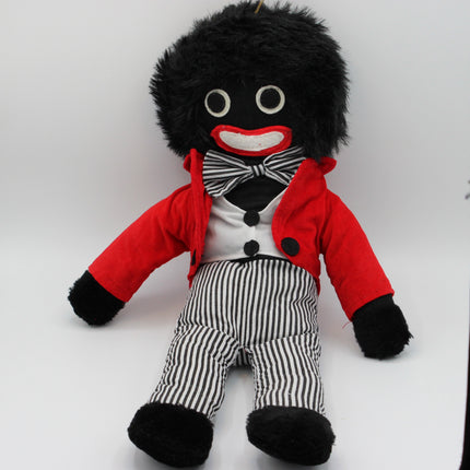 A plush Golly doll with black fuzzy hair and black-and-white striped pants, wearing a red jacket, white shirt, black bow tie, and black shoes, photographed against a plain light background.