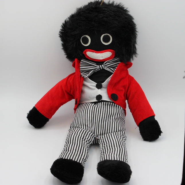 A plush Golly doll with black fuzzy hair and black-and-white striped pants, wearing a red jacket, white shirt, black bow tie, and black shoes, photographed against a plain light background.
