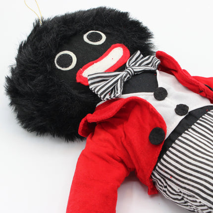 A plush  Golly doll with a black furry head, black eyes, and a wide red-and-white mouth, dressed in a red jacket, white shirt, black-and-white striped bow tie, and black-and-white striped pants on a white background.