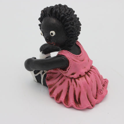 A small figurine of a seated Golly with dark skin, curly black hair, and a pink dress, photographed on a plain white background.