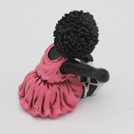 A small figurine of a seated Golly person with a large black textured hairstyle, wearing a pink dress and holding a black-and-white object, photographed on a plain white background.