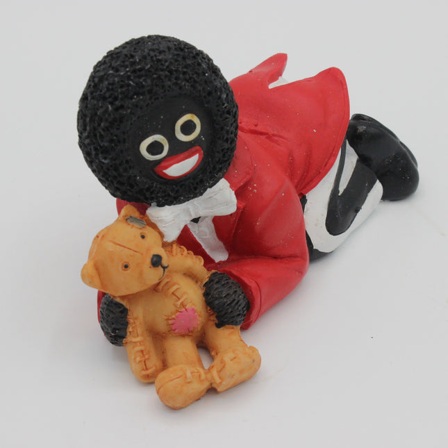 A small figurine of a black-skinned Golly character in a red outfit with a white bow, lying down and holding an orange teddy bear with a pink heart, photographed on a plain white background.