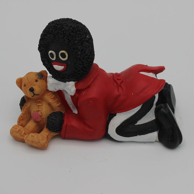 A small figurine of a black-and-white Golly character in a red coat and white bow tie reclining beside a tan teddy bear, photographed on a plain light background.