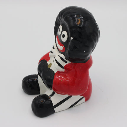 A glossy ceramic money box  figurine of a Golly character with black hair, a red top, and a white-and-black striped body, shown on a plain light background.