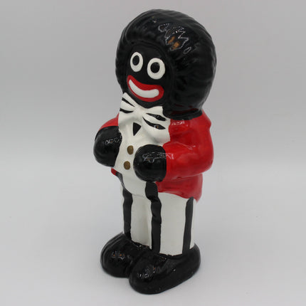 A glossy ceramic figurine of a Golly character with a black afro hairstyle, white face, red lips, wearing a red jacket, white shirt, and black pants, standing against a neutral gray background.