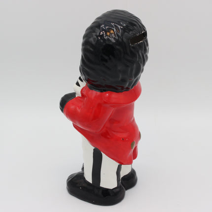 A glossy ceramic pig bank shaped like a Golly in a red coat and black-and-white striped trousers, shown from the back on a light gray background.