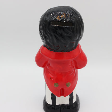 A vintage-style Golly pig bank depicting a figure from behind, wearing a bright red jacket, black hair, and a black-and-white striped lower body, set against a plain light gray background.