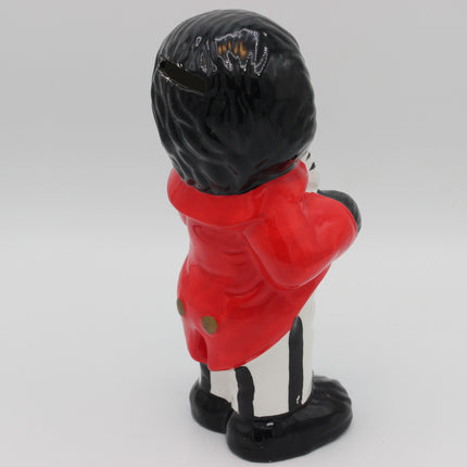 A ceramic pig bank shaped like a Golly seen from the back, wearing a bright red coat, black hair, and black-and-white striped trousers, photographed on a light gray background.
