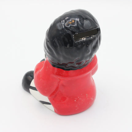 A vintage ceramic pig bank shaped like a Golly seen from behind, wearing a black hat and a bright red jacket with a white and black striped lower section, set against a plain white background.