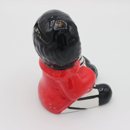 A small glossy figurine of a seated Golly viewed from the back, with a black helmet, red coat, and black-and-white striped lower body, photographed on a plain white background.